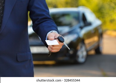 New Car And Business Theme: A Man In A Black Suit Holds The Keys Of A New Car And Business Card On A Background Of Green Tree