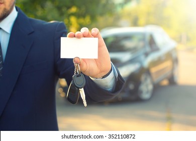 New Car And Business Theme: A Man In A Black Suit Holds The Keys Of A New Car And Business Card On A Background Of Green Tree