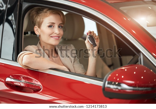 New car means\
new level in life. Attractive young blond woman smiling joyfully at\
camera and showing car key to the camera while sitting in the red\
shining car at auto\
dealership.