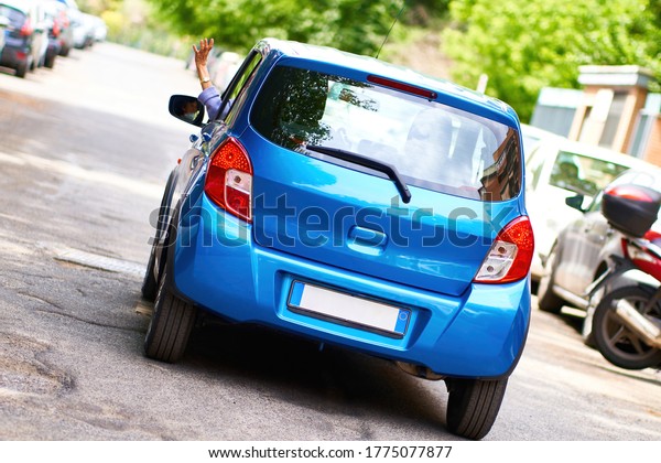 New Car: Old Lady\
Waving Goodbye, Driving Away for Holidays. Blue Car, No Logos,\
Unrecognizable Woman Wearing a Face Mask Visible in Side Mirror.\
Rome, Italy - May 28, 2020