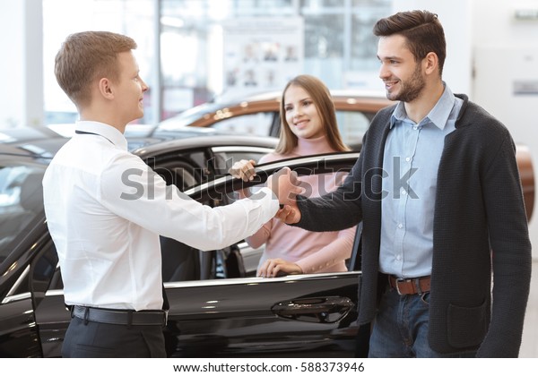 New car owners. Happy young couple buying a new\
car beautiful couple receiving car keys from a salesman at the car\
dealership lifestyle happiness family purchase owners new giving\
handing keys concept