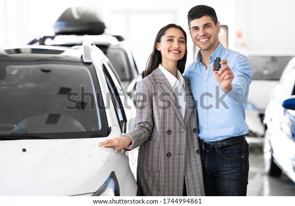 New Car Owners. Young Famile\
Standing In Dealership Store, Man Showing New Car Key, Free\
Space