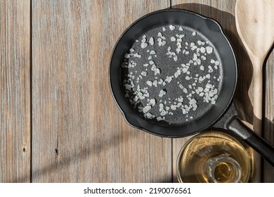 A New Clean Cast Iron Pan With Salt On A Wooden Table, A Spoon And A Bottle Of Vegetable Oil. Background With Cooking Utensils And Kitchen Utensils For Frying. Recipe Template