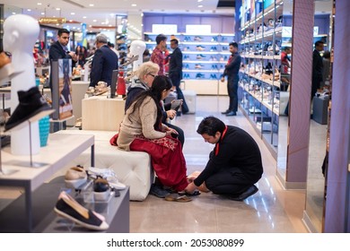 New Delhi, India- Jan 7 2020: A Woman In A Shoes Showroom, Shopping For Shoes, Sitting Down While A Male Shop Assistant Hands A Shoe To Her To Try On 