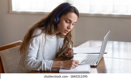 New education style. Attentive teenage girl pupil student sitting by laptop at home putting earphones on listening distant lecture remote lesson at video conference mode making notes to paper copybook