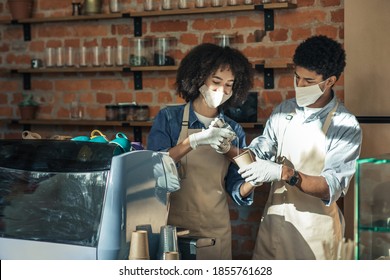 New Employee, Training And Work During Covid-19 Pandemic. Millennial African American Woman In Apron, Protective Mask And Gloves With Guy Preparing Latte Near Equipment In Interior Of Loft Cafe