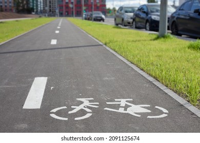 New Empty Bicycle Path Closeup With Green Grass And Cars Around. White Bike Symbol On Road. Bicycle Infrastructure In The City. Bikeway With No People. Separated Road For Cycling.