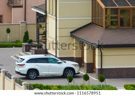 New expensive white sport car Honda parked in front of garage door in paved well-kept yard with green lawn, decorative trees and brick fence of big two story cottage. Luxury and prosperity concept.