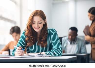 New Highschool Year On Campus. Positive Beautiful Young Adolescent Redhead Lady Sitting At Table In Modern Classroom, Doing Test Exercise, Reading Task, Enjoying Education And Learning With Classmates