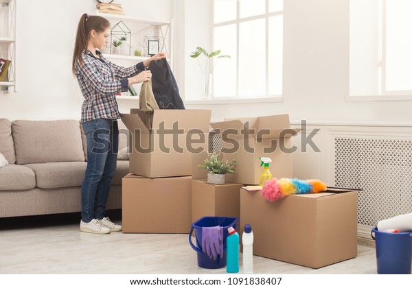 New home\
cleaning. Young woman tidying up and unpacking boxes after moving\
to new apartment. Girl with various detergents, rags and mops in\
living room full of carton\
boxes.