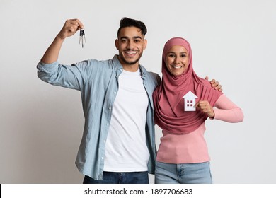 New Home Concept. Happy Muslim Couple Holding Paper House And Keys, Celebrating Buying New Property, Happy Arab Spouses Estate Owners Standing On Grey Studio Background, Lady Wearing Hijab, Copy Space