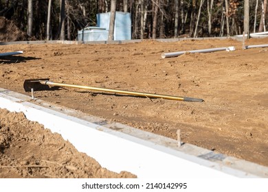 New Home Construction Site With Rake On Sand, Foundation, Pipes, And Woods.