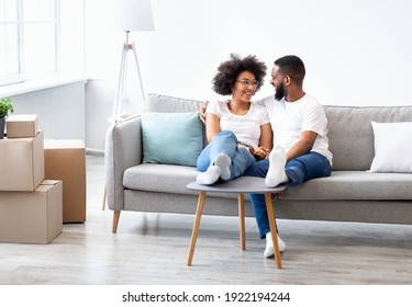 New Home. Happy Black Spouses Talking Sitting On Sofa Among Packed Boxes Enjoying Moving Day Indoors. Real Estate, House Ownership And Renting Concept. Copy Space