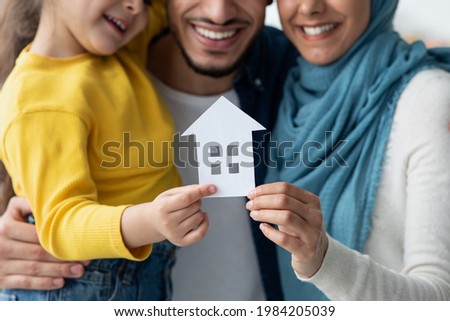 New Home. Happy muslim family of three holding cutout paper house figure in hands, arab father, little daughter and mother in hijab celebrating buying new property, real estate and insurance concept