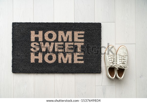 New home moving in door mat\
entrance welcome doormat with text writing HOME SWEET HOME and\
white sneakers of happy condo homeowner. Top view of rug and wooden\
floor.