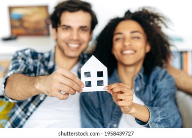 New Home Owners. Joyful Multiethnic Couple Holding Paper House, Sitting On A Sofa In Their New Modern Apartment Or House. Defocused African American Girl And Hispanic Guy Happy To Buy Their Own Home