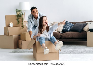 New Home, Relocation. Happy Mixed Race Young Family Couple, Caucasian Girl And Latino Guy, Is Playing With A Cardboard Box, Girl Sits Inside And Guy Push It, Their Have Fun Together At New Apartment