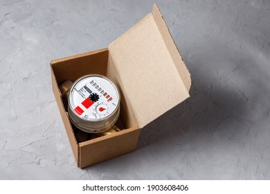 A New Hot Water Meter In A Cardboard Box On A Gray Concrete Background.