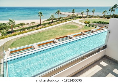 New Hotel Balcony With With Glass Fencing Over Large Swimming Pool Against Blue Sea On Clear Horizon On Sunny Day In Greece