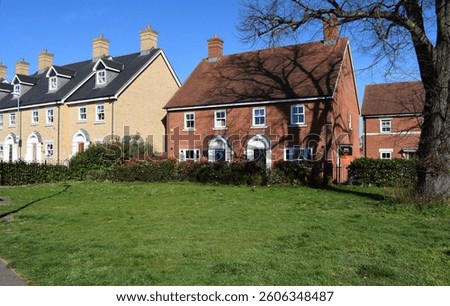A new housing development features red-brick semi-detached houses and buff-coloured brick terraced homes with traditional architectural details, adorned by lush green lawns in front and an old tree 