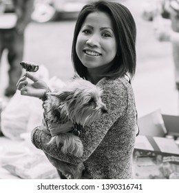 NEW IBERIA, L.A. / USA - FEBRUARY 28, 2015: A Cambodian, Kampuchean Woman Eating Holding A Jalapeño Popper & A Yorkshire Terrier In Her Arms At A Birthday Party In South Louisiana. Black And White.