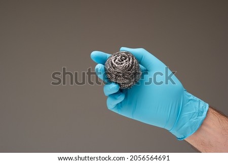 New kitchen metal wire scrubber held in hand by Caucasian male hand wearing a blue latex glove. Close up studio shot, isolated on gray background.