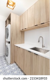 A New Laundry Room With Wood Cabinets, A White Marble Countertop, Patterned Tile Floor, White Washer And Dryer, And A Gold Light Fixture.