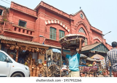 New Market, Kolkata, 03-27-2021: Low Angle View Of A Middle Aged Manual Labour Carrying Basket Filled Goods On His Head, Walking Though Congested Street Near The Iconic Hog Market, A Heritage Building