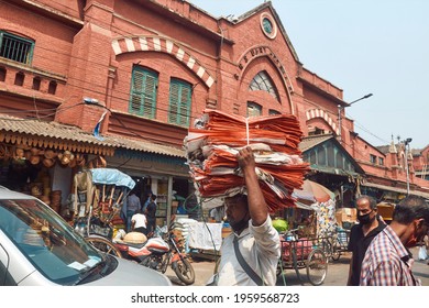 New Market, Kolkata, 03-27-2021: Low Angle View Of A Middle Aged Manual Labour Carrying Basket Filled Goods On His Head, Walking Though Congested Street Near The Iconic Hog Market, A Heritage Building