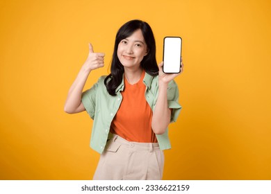 new mobile application with a cheerful young Asian woman 30s, donning orange shirt and green jumper, displaying smartphone screen with a thumbs-up gesture on yellow studio background. - Powered by Shutterstock