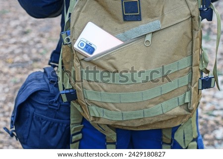 a new mobile phone with a glass camera in the unzipped back pocket of a man's military-colored khaki backpack on the shoulders of a tourist during the day on the street