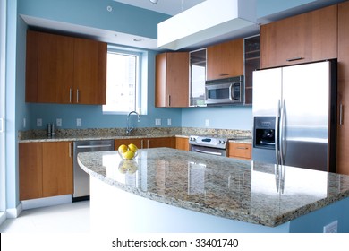 New Modern Kitchen Interior With Island In A Condo Apartment. Brightly Lit, Light Blue Walls, Granite Counter Tops, Stainless Steel Appliances. A Bowl Of Lemons On The Counter Top.