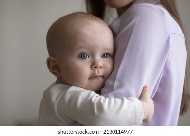 New mom holding innocent baby in arms, hugging child with love, care, affection. Adorable blue eyed infant kid embracing mother, holding shoulder, looking away. Motherhood, childbirth, family concept