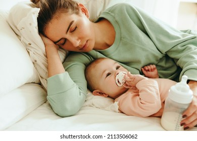 New Mom Taking A Nap With A Bottle Of Formula Milk In Her Hand. Caring Mom Resting While Her Baby Sucks On A Pacifier. Mother And Baby Lying On The Bed Together.