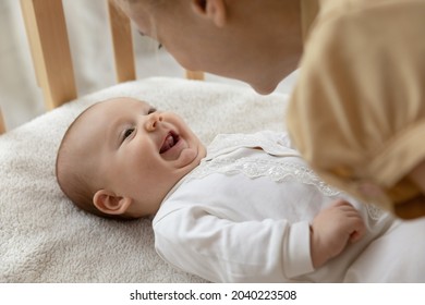 New mom talking to happy adorable few month baby resting in crib and smiling. Young mother speaking and playing with infant son or daughter lying on back. Child care, motherhood concept