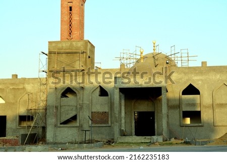 A new mosque under construction against the sunny blue sky with the mosque minaret and the domes surrounded with wood scaffold and windows under construction, the mosque is yet a cement from outside