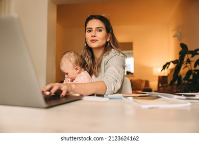 New mother typing on a laptop while working in her home office. Multitasking mom working on a new creative project at her desk. Female interior designer carrying her baby on her laps. - Powered by Shutterstock