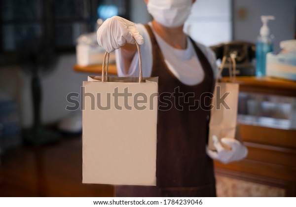 New normal An asian woman wearing gloves and medical\
face masks delivering take away food bags to customers at the\
restaurant bar to prevent the spread of corona virus.takeaway\
concept. space for logo
