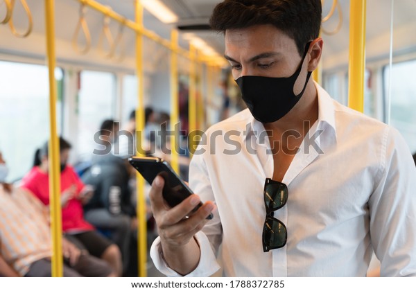 New normal attentive Caucasian man wearing face\
mask in the public metro during coronavirus pandemic, A man wearing\
face shield and casual white shirt using smartphone while business\
travel.