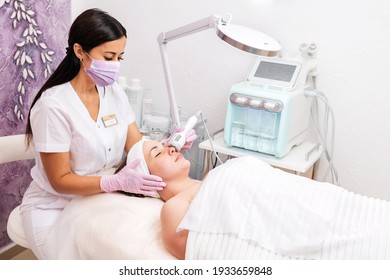 New normal. A cosmetologist in rubber gloves and medical mask does a rf-lifting procedure on the face. In the background, the Rf cosmetology device. Concept of professional cosmetology and health.