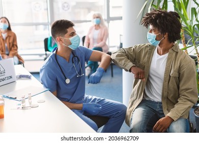New Normal Elbow Bump Greeting. Young Afro Guy In Medical Mask Bumping Elbows Meeting Male Doctor In Hospital Interior. Social Distancing And Keeping Safe Distance For Covid-19 Protection Concept