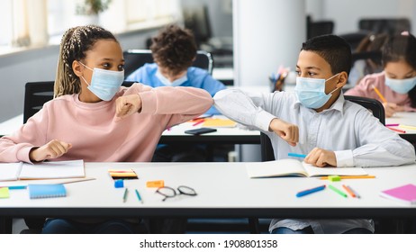 New Normal Greeting. Diverse multiethnic schoolchildren doing elbow bumping sitting together at table and wearing disposable surgical face masks. Black girl and boy avoid coronavirus spread - Powered by Shutterstock