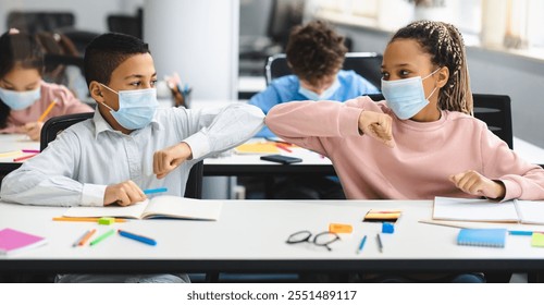 New Normal Greeting. Diverse multiethnic schoolchildren doing elbow bumping sitting together at table and wearing disposable surgical face masks. Black girl and boy avoid coronavirus spread - Powered by Shutterstock