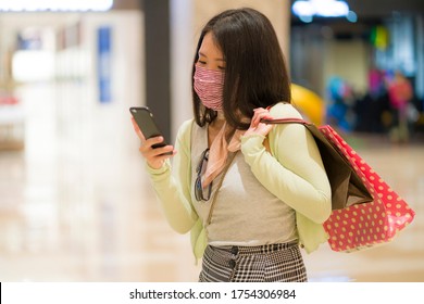 New Normal Habits - Young Attractive And Happy Asian Chinese Woman Using Mobile Phone At Mall Happy And Excited Enjoying Sale In Beauty Fashion Store Carrying Shopping Bags 