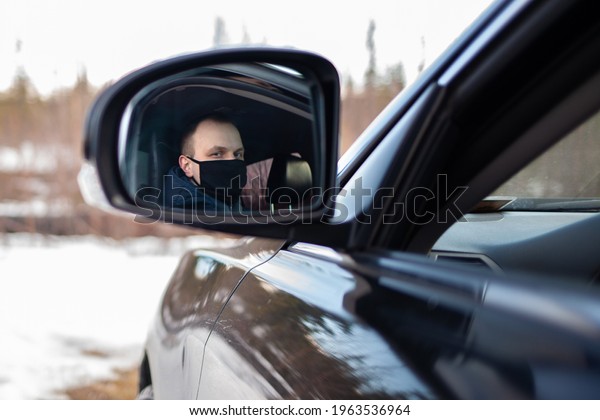 new normal man,\
reflection of young male driver in black medical mask in rearview\
mirror sitting in black\
car.