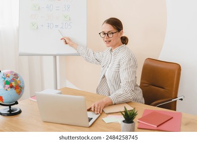 New normal. Maths distant remote online lesson. Successful young teacher tutor lector working on distance during lockdown, explaining sums on whiteboard using laptop for video call.