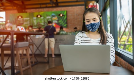 New Normal And Social Distancing After Covid Epidemic Concept.young Smart Asian Female Watching Laptop Meeting Business Team Or Webinar Course Online With Face Shield Or Mask Protection At Coffee Shop