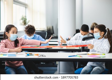 New Normal Social Distancing. Diverse Pupils Wearing Protective Face Masks Keeping Distance At Arm's Length, Sitting At Desks In Classroom With Outstreched Arms. Stop Spreading Virus