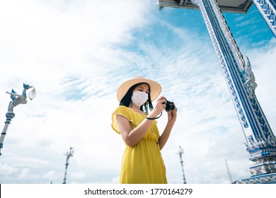 New Normal Travel Lifestyle After Virus Covid Concept. Adult Asian Woman Wear Mask And Yellow Dress Hold Camera For Take Photo. Outdoor Local Wanderlust At Temple In Asia On Day. Chantaburi, Thailand