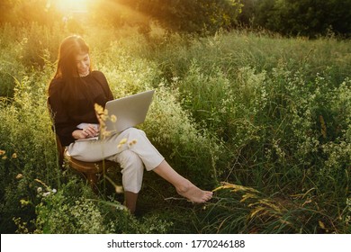 New Office Concept. Fashionable Elegant Girl Working On Laptop And Sitting On Rustic Chair In Warm Sunset Light In Summer Field. Remote Work With Social Distancing And Safety Protocols